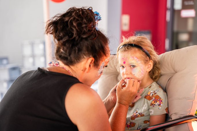 Frau schminkt ein Kind mit bunten Gesichtsmalerei in einem fröhlichen Raum.
